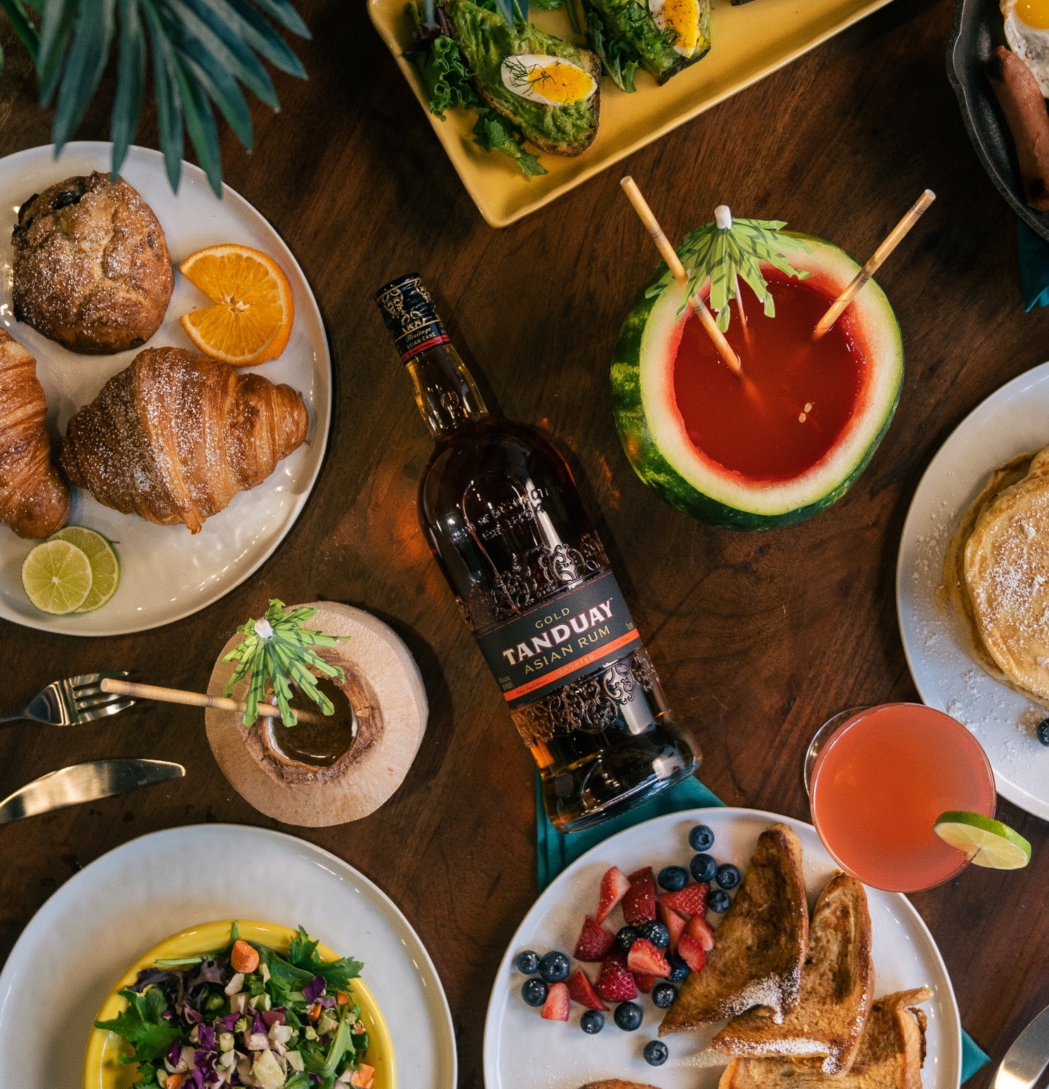 Пляшка рому Tanduay Asian Rum Gold на святковому столі з десертами, фруктами й тропічними коктейлями