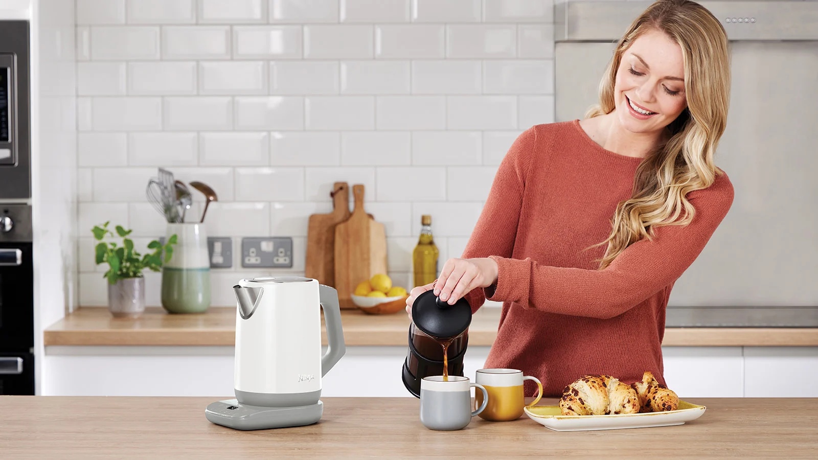 Жінка на кухні наливає каву з френч-пресу в чашку, поруч електрочайник Ninja і тарілка з круасанами