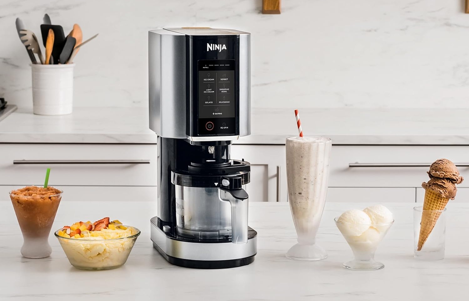 Морозивниця Ninja на кухонному столі з приготованими десертами — морозивом, сорбетом, мілкшейком і фруктовим боулом.
