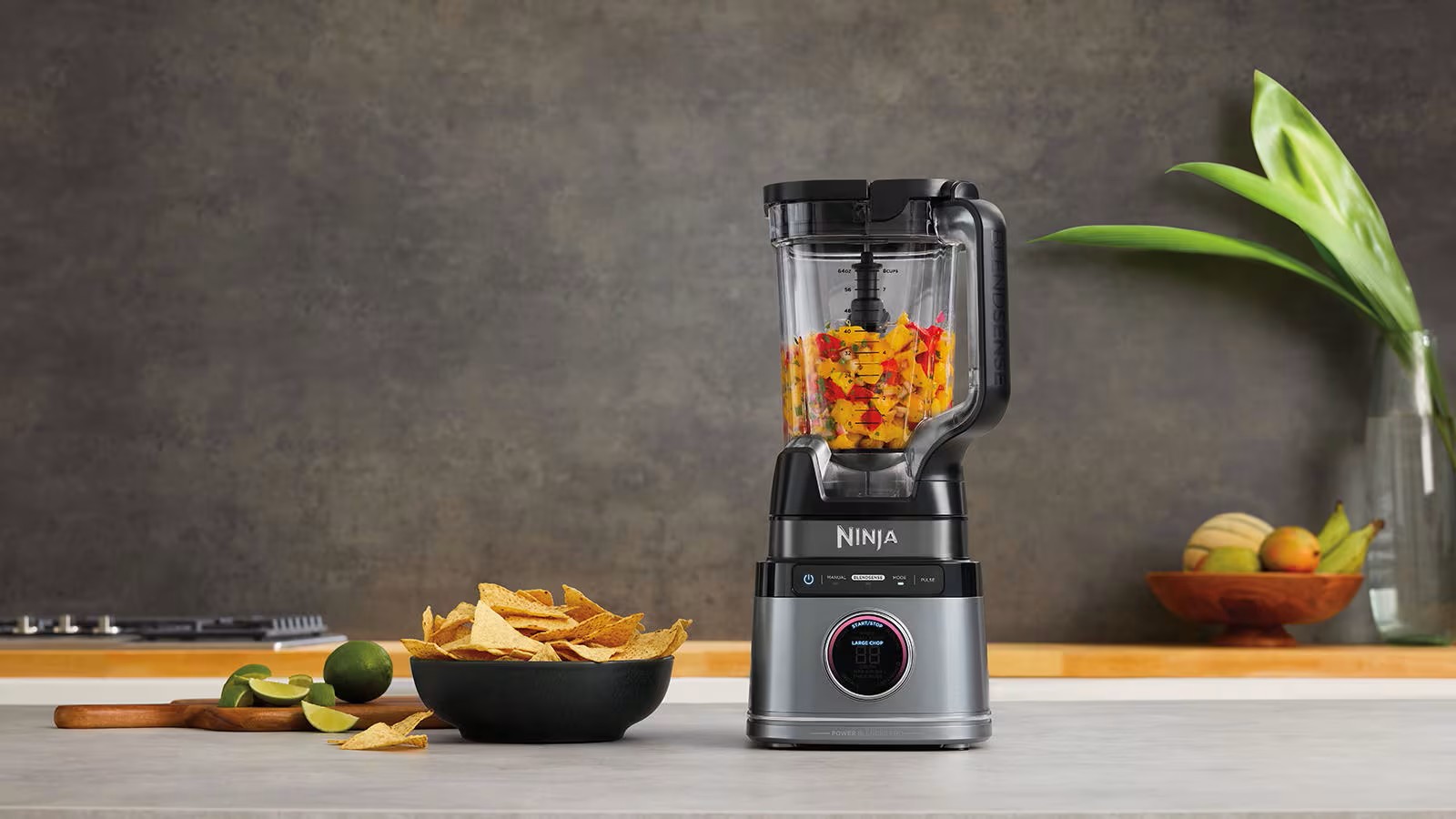 Стаціонарний блендер Ninja з подрібненими овочами всередині чаші, поруч миска з чипсами та лайм