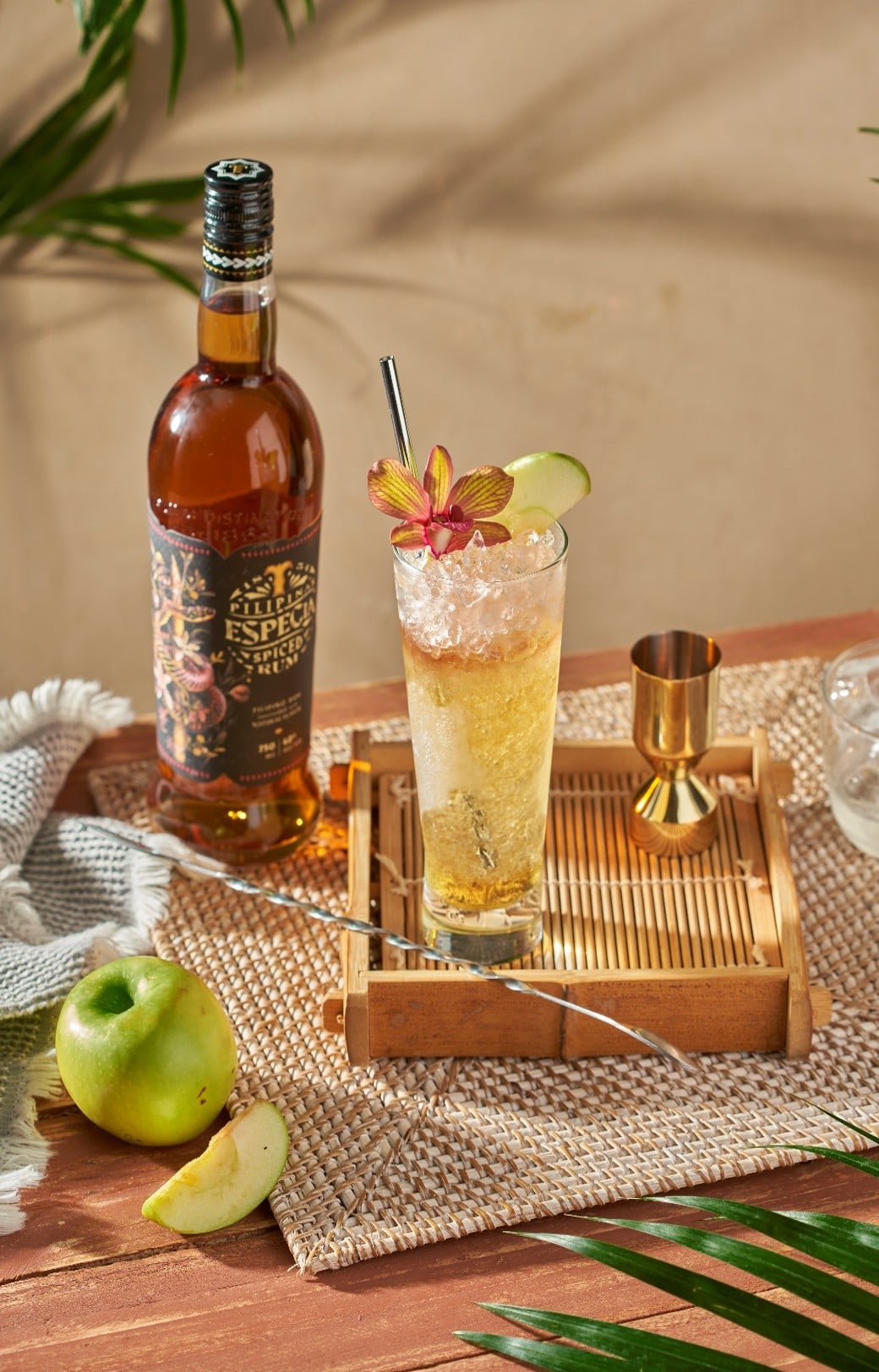 Пляшка рому Especia Spiced Rum поруч із коктейлем із льодом, яблуком і квіткою на плетеній серветці з тропічними акцентами