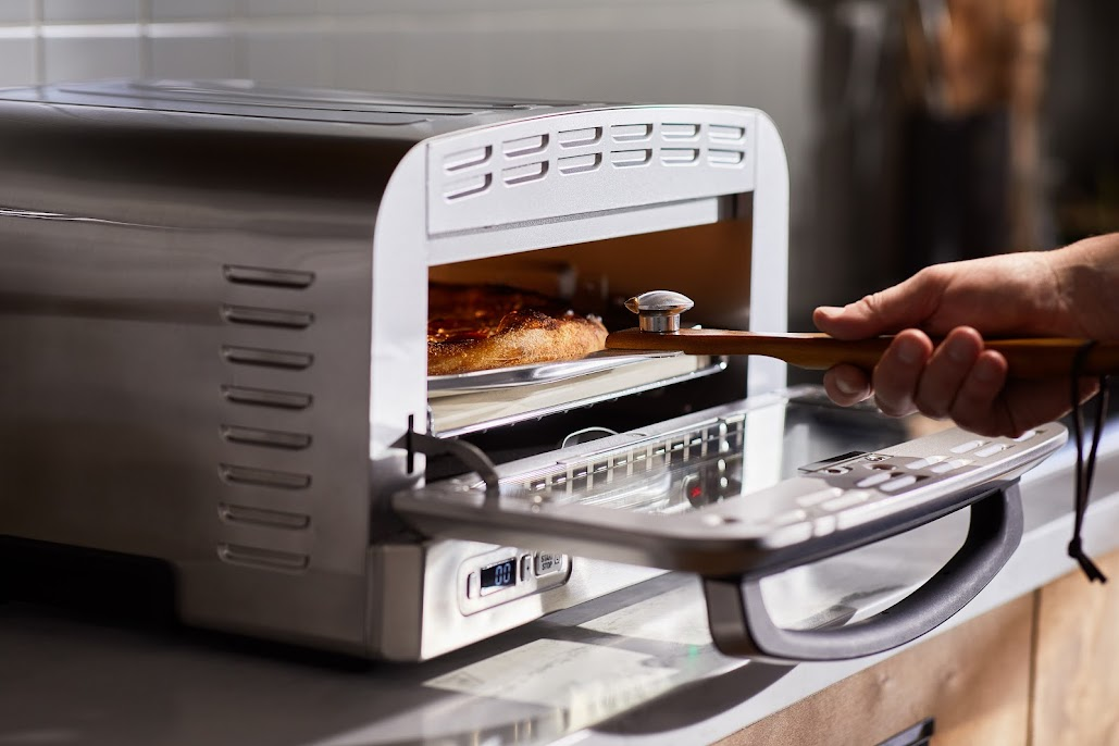 Процес приготування піци в електричній печі Cuisinart, рука тримає лопатку для піци біля відчинених дверцят печі