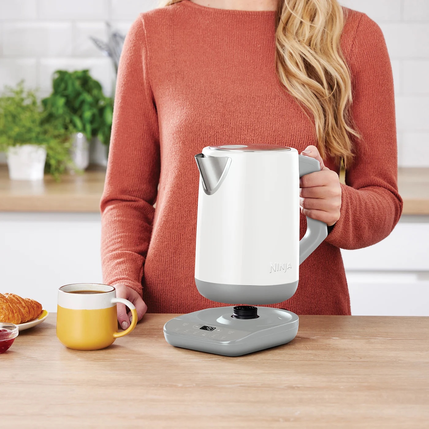 Жінка тримає електрочайник Ninja білого кольору на кухні, поруч чашка з гарячим напоєм і круасан