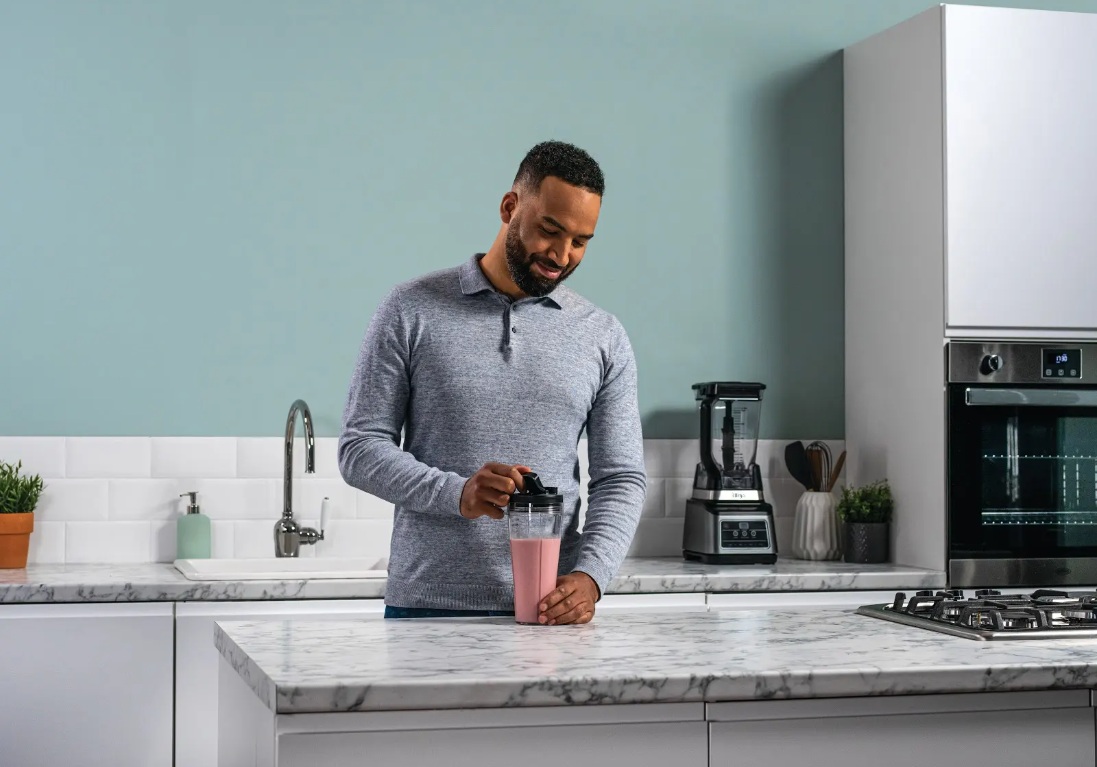 Чоловік стоїть на кухні та тримає склянку з рожевим смузі, на задньому плані стоїть блендер Ninja