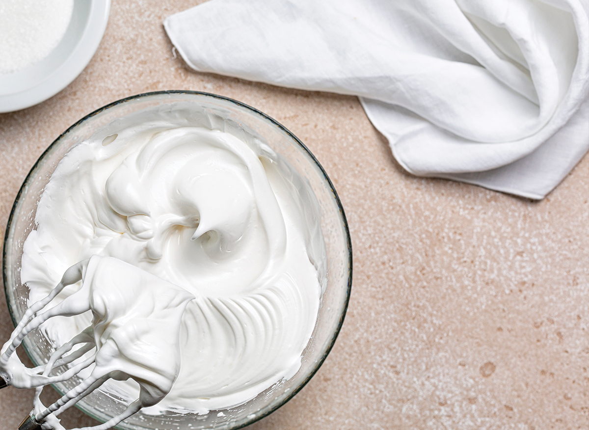 Збита до пишних піків білкова маса в скляній мисці поруч із вінчиками міксера