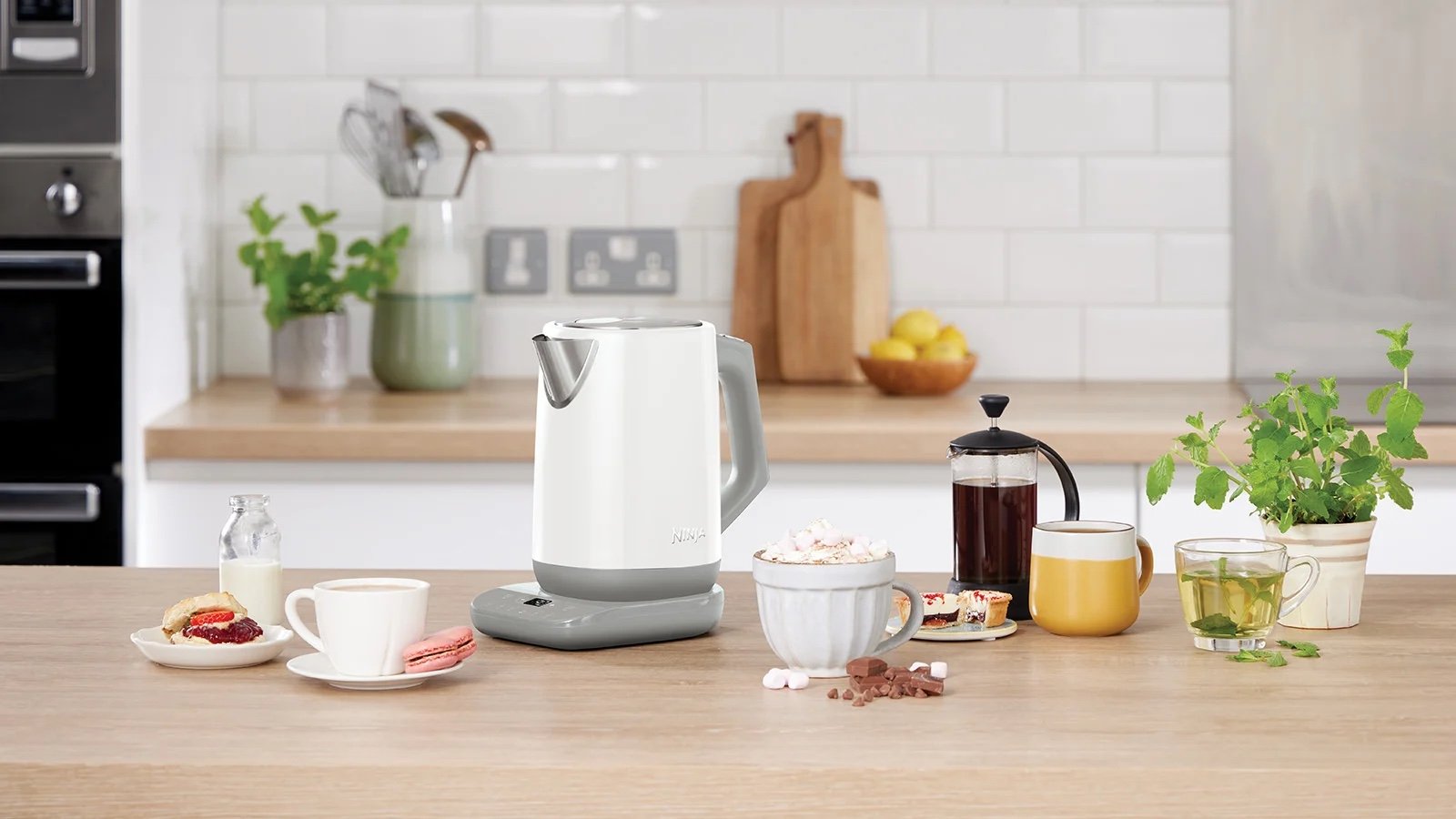 Електрочайник Ninja на кухонному столі поруч із чашками чаю, кавою, десертами та свіжою м’ятою