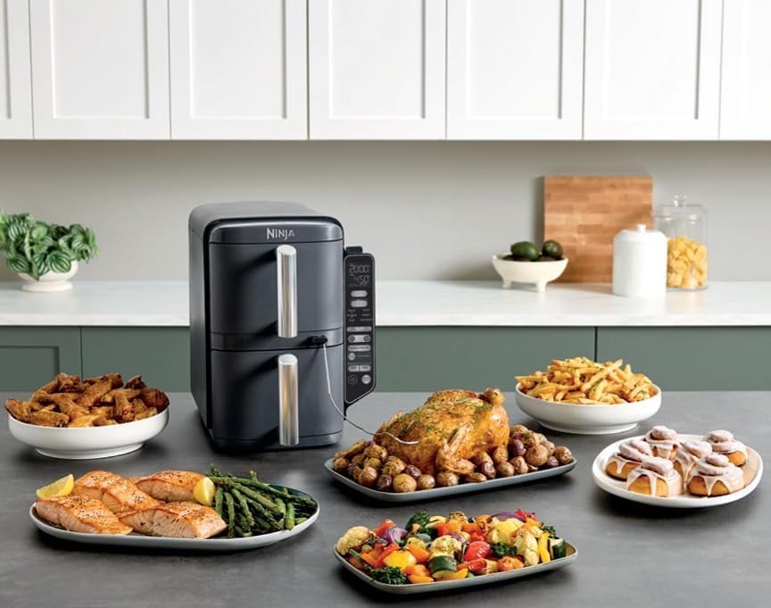 Ninja Double Stack на кухонному столі поруч із готовими стравами — куркою, овочами, картоплею фрі, рибою та десертами.